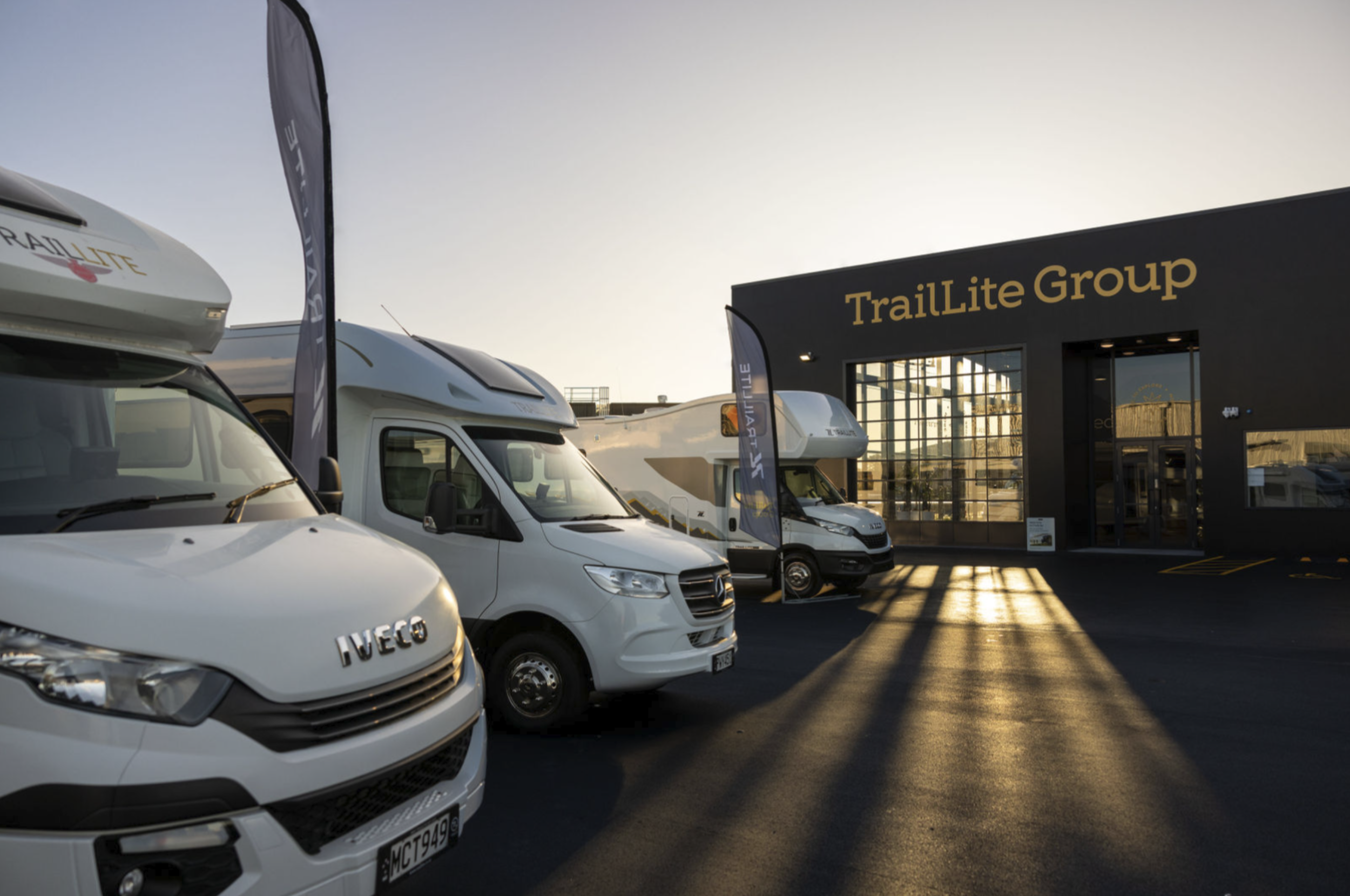 Multiple recreational vehicles parked outside a black building with a TrailLite Group sign during sunset, with sunlight casting long shadows